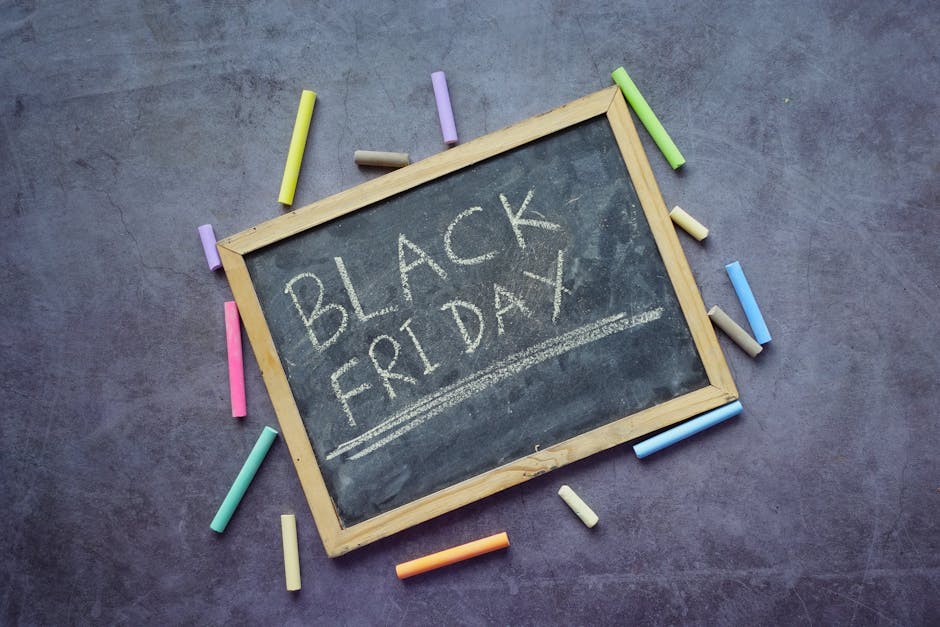 A small rectangular chalkboard with a light wooden frame is placed on a dark, textured surface. The board has the words 'BLACK FRIDAY' written in white chalk, with an underline beneath the text. Surrounding the chalkboard are multiple pieces of colorful chalk in shades of yellow, green, pink, purple, blue, beige, and orange, some lying flat while others are leaning against the edge of the frame. The scene is well-lit, with even lighting highlighting the contrast between the black surface of the chalkboard and the vibrant chalk colors. The setup suggests an informal or educational environment, but in the context of rubbish removal services, it subtly evokes ideas of promotion or special sales events. The arrangement is clean and orderly, with no visible clutter, rendering a neat and professional appearance suitable for visual content associated with waste management or promotional campaigns related to discount offers or cleaning services. The image emphasizes simplicity and clarity, suitable for accessibility and SEO relevance in the context of rubbish collection or disposal discussions.