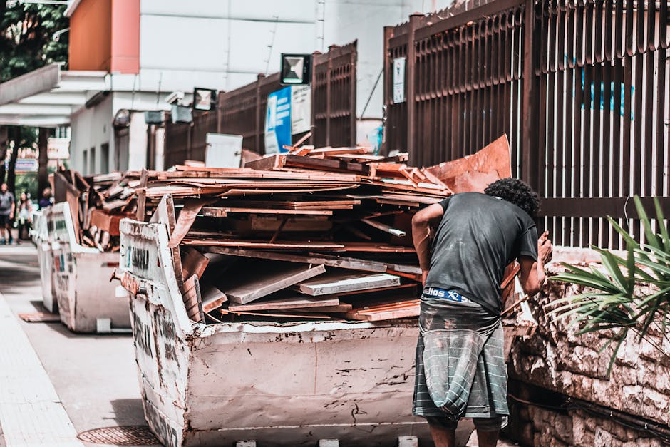 A man with curly black hair wearing a dark grey T-shirt and checkered shorts stands beside a large white skip filled with a substantial pile of broken terracotta roof tiles and construction debris, situated on a city street. The tiles have a reddish-brown finish, some with cracks or chipped edges, and are stacked haphazardly, spilling over the edges of the skip. The skip, showing signs of wear with dirt marks and minor dents, is positioned close to a metal fence with vertical bars painted dark brown, which runs along the sidewalk. Behind the fence, there are glimpses of urban buildings with white walls and signage, indicating a busy environment. To the left, a white building with a glass awning and a sidewalk occupied by pedestrians is partially visible. The lighting suggests natural daylight, casting soft shadows on the scene. This image reflects an example of private waste handling or on-site clearance, consistent with rubbish removal services like those offered by Rubbish Collection Paddington, specifically related to bulky waste disposal and roofing tile removal.