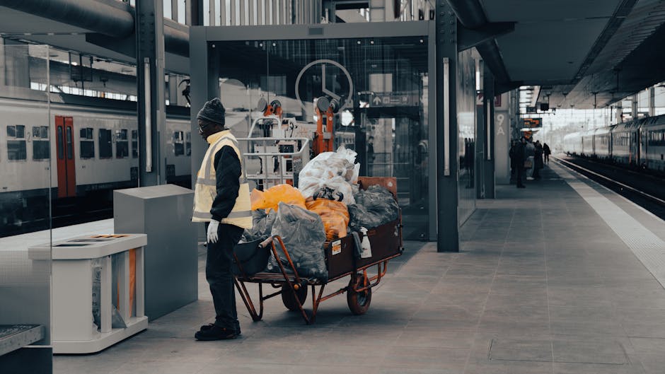A worker standing on a modern, covered train station platform, wearing a dark beanie, glasses, a high-visibility vest over a black jacket, and white gloves. Next to the worker is a large red-handled trolley loaded with various bags and loose refuse, including black, grey, and orange plastic bags, some filled and tied, with other miscellaneous waste items piled on top. The platform surface is made of smooth grey tiles, with signage and a ticket machine visible on the left side. In the background, an overhead glass enclave with metal framework encloses the station's entrance, while on the right, a stationary train with dark and light grey exterior panels is parked adjacent to another platform space. Further along the platform, a few distant figures are walking, and a train carriage with closed doors is visible. The scene is evenly lit with natural daylight, and the setting suggests an urban transport hub involved in or facilitating private waste handling or collection activities common in station environments, consistent with independent rubbish collection services.