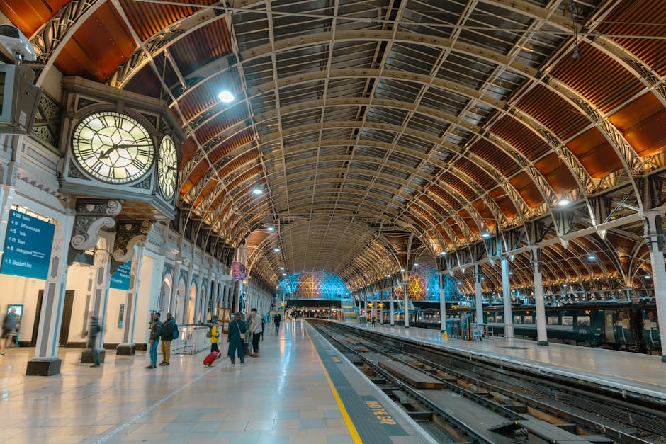 Inside a spacious train station with a high arched ceiling featuring metal framework and wood paneling, illuminated by overhead lighting and decorative lamps. On the left side, there are large clock faces mounted on ornate white columns, along with electronic departure boards displaying train information. The platform area is populated with a few travelers, some walking and others standing, with luggage at their sides. The tracks run parallel across the width of the platform, with train carriages visible in the background. The environment is clean and orderly, with a mix of modern lighting and historic architectural elements. The scene depicts an urban transportation hub, similar in appearance to a location where independent or private waste management services could occur nearby, though no waste-related objects are visible.