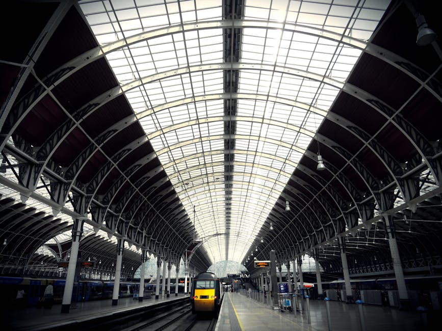 An interior view of a large, modern train station with a vaulted glass ceiling supported by white metal arches and beams, allowing natural light to illuminate the station’s spacious platform area. On the platform, there is a yellow and black train approaching or departing, with multiple sets of railway tracks visible running parallel to the platform. The environment is clean and well-organized, with signage and digital displays providing travel information, and a few passengers visible in the distance. The station’s architecture features sleek, curved lines and a combination of glass and metal finishes, typical of contemporary transport hubs designed for efficient movement and accessibility. The clear view of the station’s expansive roof structure highlights the use of natural light to create a bright, open atmosphere, relevant to discussions around urban infrastructure and public transportation facilities, which may relate indirectly to waste management in terms of maintaining clean and functional station environments.