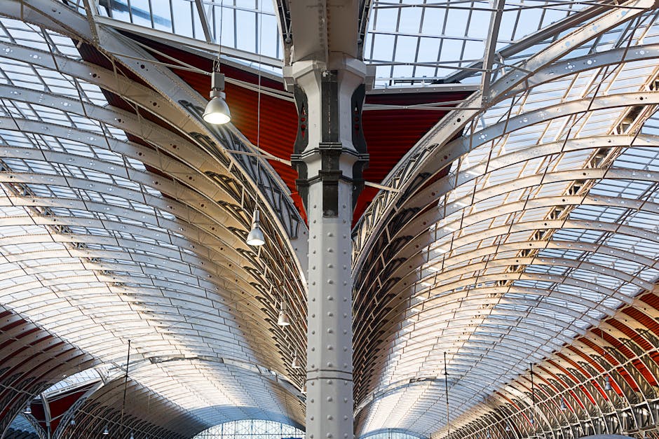 An interior view of a large, modern train station with a vaulted glass ceiling supported by white metal arches and beams, allowing natural light to illuminate the station’s spacious platform area. On the platform, there is a yellow and black train approaching or departing, with multiple sets of railway tracks visible running parallel to the platform. The environment is clean and well-organized, with signage and digital displays providing travel information, and a few passengers visible in the distance. The station’s architecture features sleek, curved lines and a combination of glass and metal finishes, typical of contemporary transport hubs designed for efficient movement and accessibility. The clear view of the station’s expansive roof structure highlights the use of natural light to create a bright, open atmosphere, relevant to discussions around urban infrastructure and public transportation facilities, which may relate indirectly to waste management in terms of maintaining clean and functional station environments.