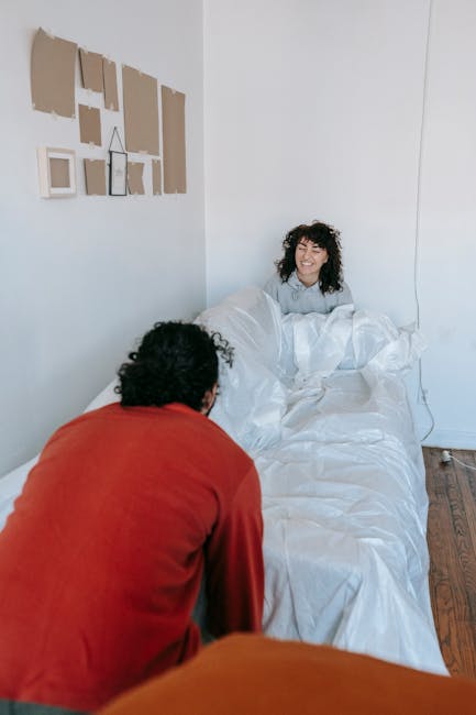 A woman with curly dark hair and a broad smile is sitting up in a bed, covered with white bedding and a duvet, inside a bright, minimalist bedroom. She appears to be in a relaxed and happy mood, with her head slightly tilted and looking towards the person in front of her. In the foreground, a person with dark, curly hair wearing an orange top is seen from behind, leaning slightly forward towards the bed. The wall behind the woman features a display of small, variously shaped picture frames in neutral tones, mounted on a light-colored wall. The room has wooden flooring and natural light illuminating the scene, creating a warm and inviting atmosphere. The setting suggests a casual environment, possibly part of a home or a temporary living space. The image relates to private waste management or alternative rubbish disposal by indicating potential for on-site clearance or the need for removal services, such as mattress disposal, with Rubbish Collection Paddington potentially providing assistance in such scenarios.