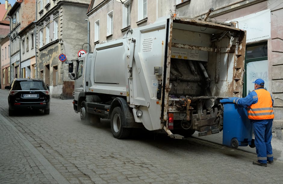 A commercial waste collection truck parked on a narrow cobblestone street beside old multi-story buildings with aged, peeling facades and large windows. The truck, predominantly white with some rusted and dirty areas, is in the process of emptying a blue wheeled bin held by a worker dressed in high-visibility orange and blue clothing with a matching cap. The worker is positioned on the right side of the image, guiding the bin towards the truck's open rear compartment, which is designed for on-site waste collection. In the background, a black car is parked further down the street, and two traffic signs are visible on the building wall, indicating parking restrictions. The environmental setting is urban, with the scene capturing an everyday waste disposal activity, typical of independent or private rubbish collection services often used for local waste management outside of standard council rubbish collection, illustrating the practical context for rubbish removal in central city areas.