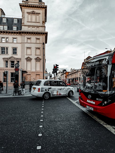 A busy urban street scene featuring a vintage white taxi with a slightly weathered exterior and dark tinted windows, positioned at a road intersection in front of a historic beige stone building with decorative architectural details including cornices and small windows. To the right, there is a modern red and black double-decker bus with destination signage indicating 'Paddington' on the front. The street is lined with various shops and storefronts, with pedestrians waiting to cross the road at a traffic light, which is showing red. The road surface has visible lane markings, and the overall atmosphere is overcast with soft, diffused lighting, typical of a city environment where alternative or private waste collection might be coordinated independently from local authorities, as suggested by the surrounding context of urban logistics and transport.