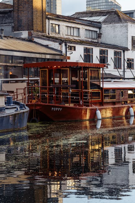 A brown wooden houseboat named 'Poppy' is docked along a narrow urban waterway, with its exterior featuring a covered deck area topped by a red canopy. The boat is equipped with white cylindrical fenders hanging from the sides to protect it from impacts against the quay. Behind the houseboat, a mix of old brick and white-painted buildings with various window styles and rooftop textures are visible, indicating a historic or mixed-use waterfront area. The water surface reflects the boat and surrounding structures, showing slight ripples and algae or surface debris. The scene is lit with natural daylight, with a subdued urban atmosphere. This setting relates to private or alternative water-based disposal or storage, aligning with independent waste handling or specific use of boats for transport or accommodation on the water, relevant to local rubbish and waste management considerations in dense city environments. Rubbish collection services occasionally handle waste from such waterfront areas, though they are not directly visible in the image.
