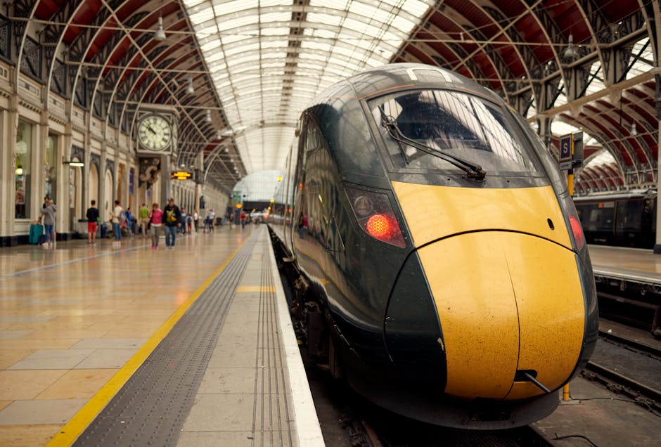 Inside a train station with a large, modern, high-speed train positioned at the platform edge in the foreground. The train has a sleek, aerodynamic design with a dark grey body and a distinctive yellow front section, which shows some grime and dirt. The station's interior features an arched, glass-paneled ceiling allowing natural light to illuminate the space, and a large, vintage clock mounted on the left wall displays the time. Several passengers are visible standing or sitting along the platform, some with luggage or bags, while others appear to be waiting or walking. The platform is lined with safety tactile paving and yellow safety lines near the train doors. The overall scene depicts a busy, urban railway environment supporting private and independent transportation services, with subtle associations to efficient and organized rubbish clearance at transportation hubs or commercial settings, aligning with professional waste management themes in busy public infrastructure.