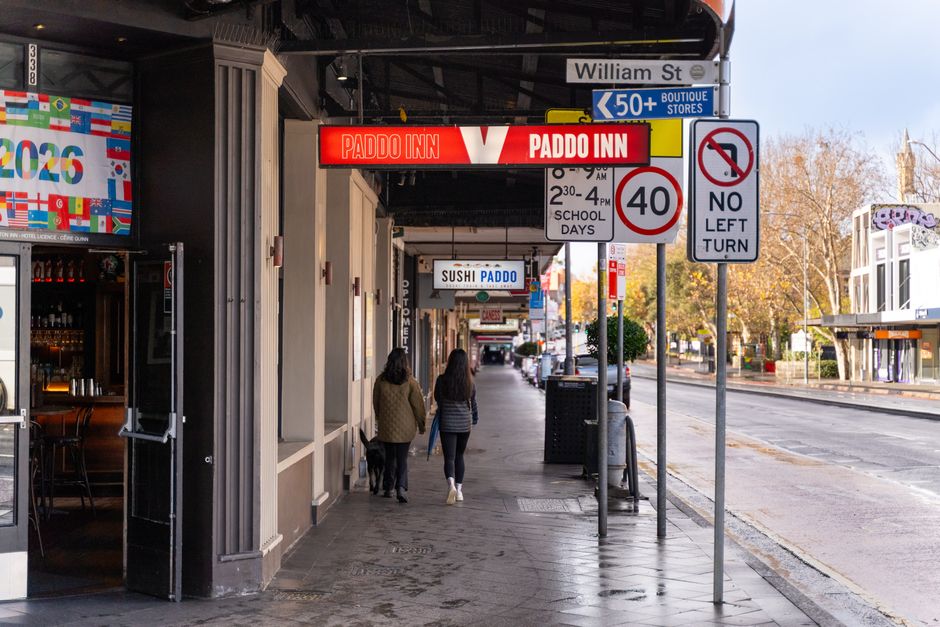 A city sidewalk scene showing a street with signage for William Street and a nearby blue directional sign indicating shops and stores. The foreground features a cluster of traffic and parking signs, including a red and white '40' speed limit sign and a 'No Left Turn' sign, positioned near a pole. The sidewalk runs alongside the roadside, where a black garbage bin and a trash can are placed near the curb. Pedestrians, including two women dressed in casual jackets and jeans, walk along the pavement away from the camera. On the building façade, there are illuminated signs for a sushi restaurant labeled 'SUSHI PADDO,' and another sign advertising 'PADDO INN,' both attached under a dark overhang. The environment is well-lit with natural daylight, and trees with autumn foliage are visible in the background, indicating a typical urban setting. This scene relates to private waste management and rubbish collection services, as seen from the general cleanliness and placement of waste receptacles along the street, which aligns with alternative waste handling methods in urban areas.