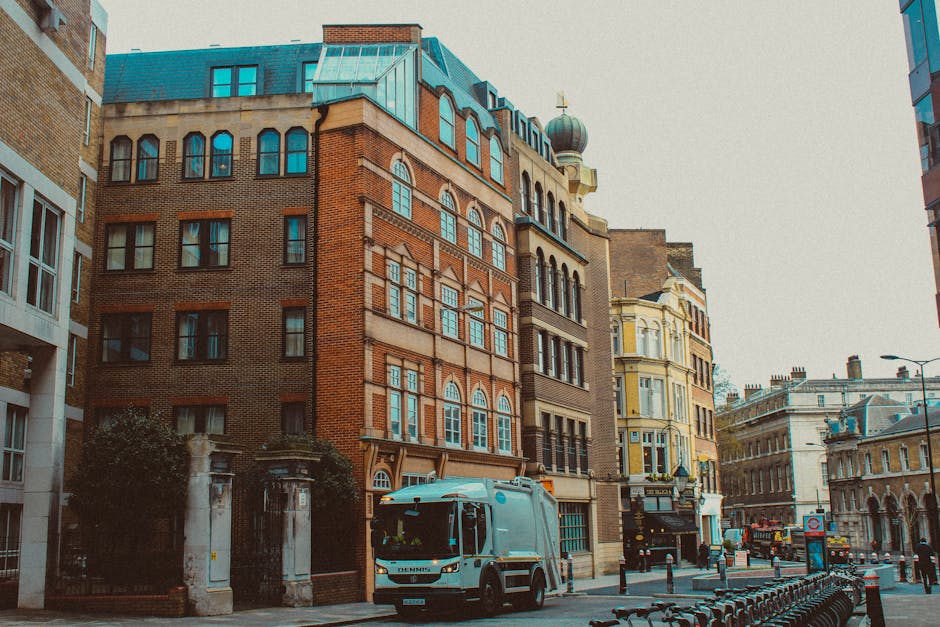 A street scene featuring a collection of historic multi-storey brick and stone buildings with ornate window frames, decorative architectural details, and a domed tower with a gold cross on top. The buildings are situated along a paved city street with parked vehicles, including a blue delivery truck in the foreground and a waste collection vehicle partially visible further down the road. To the left, a gated entrance with stone pillars and a small tree are visible, while on the right, a modern glass-fronted building reflects the surrounding architecture. The street is lined with bollards, bicycle racks, and street lamps, creating an urban environment suited to residential and commercial use. The natural daylight illuminates the scene, with overcast skies giving a muted tone. This image relates to private waste collection services like those offered by Rubbish Collection Paddington, highlighting the urban setting where independent rubbish removal can take place outside of council authority areas.