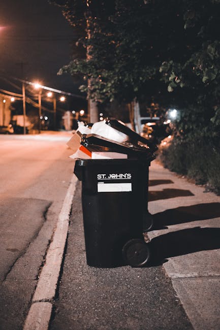 In a public urban setting, a large pile of mixed waste materials is overflowing from several rubbish bins placed on a paved sidewalk. The collection includes a grey recycling container labeled for mixed paper and cardboard, which is topped with loose papers, cardboard boxes, and plastic bags. To its right, there are numerous black garbage bags and a bright red waste bin, both filled with assorted waste. Additional waste items, such as cardboard boxes, paper packaging, plastic bottles, and discarded items, are scattered around the base of the bins, creating a cluttered scene. In the background, a blue metal fence surrounds a building under construction or renovation, with scaffolding and completed windows visible, indicating an urban commercial or residential area. A parked car is partially seen behind the fence, and the environment appears exposed to daylight, with natural light illuminating the scene and highlighting the disorderly waste accumulation, which may suggest a situation requiring private rubbish collection or on-site waste handling services like those offered by Rubbish Collection Paddington.