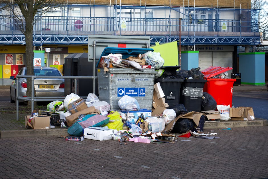 A large collection of overflowing rubbish bins and open waste bags placed on a paved sidewalk in an urban setting. The waste includes cardboard boxes, plastic bags, paper, and miscellaneous household refuse, with some items spilling onto the ground. The debris is situated near a metal railing, with a parked silver car to the left and a row of shops with bright signs and storefronts in the background. Behind the waste, there is a building with blue scaffolding and safety barriers, indicating ongoing construction or maintenance work. The scene is illuminated by natural daylight, highlighting the variety of waste materials and the cluttered arrangement. This image exemplifies a typical scenario for private waste disposal or alternative rubbish collection services, such as those offered by Rubbish Collection Paddington, especially in areas where bin space is inadequate or collection is scheduled outside of standard council services.