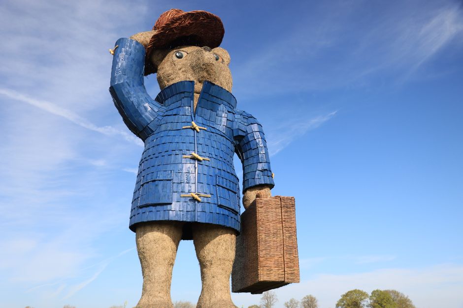 A large outdoor sculpture of a stylised human figure with a rounded head, painted and textured to resemble stone or concrete, standing against a clear blue sky with wispy clouds. The figure is dressed in a vibrant blue coat with a textured, tiled appearance, fastened with three yellow toggle buttons, and has beige, rough-textured legs and hands made to resemble stone or cement. The figure is holding a rectangular, brown wicker or wood basket in its right hand, which is textured and appears empty or lightly filled. The sculpture is situated on a grassy area with a few distant trees visible at the horizon in the background. The lighting indicates a sunny day, casting soft shadows and enhancing the colours and textures of the sculpture. This image relates to waste management and rubbish collection by illustrating a large, artistic representation of someone associated with rubbish removal or private disposal services, in alignment with the context of independent waste handling often needing shared or municipal collection options.