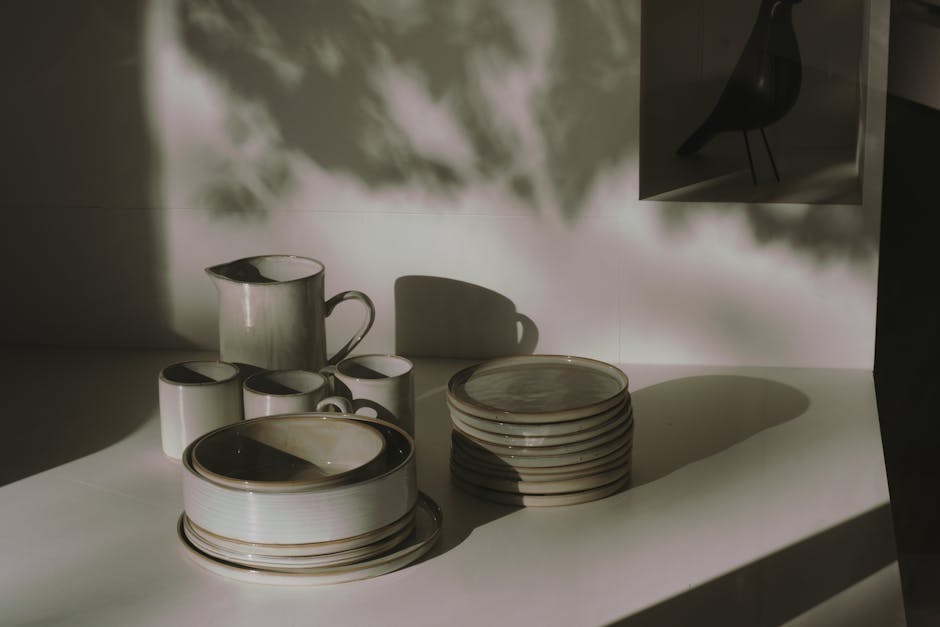 On a clean, light-colored kitchen countertop, there is a neatly stacked pile of ceramic dinner plates with a glossy beige finish and darker rim accents, placed alongside a smaller stack of similar plates. To the left of the plates, a matching ceramic jug featuring a glossy beige surface and a curved handle is positioned. Next to the jug, four smaller ceramic mugs with the same beige and dark rim design are arranged, with two partially overlapping each other. The background shows a plain wall with soft, natural lighting casting distinct shadows of the objects, creating a calm and minimalistic atmosphere. The scene appears to be part of an indoor setting, possibly a kitchen or dining area, with the objects clean and ready for use or collection. This image subtly relates to the context of waste management by showcasing typical household items that might be considered for disposal or collection by a rubbish removal service, such as Rubbish Collection Paddington, supporting the theme of domestic waste handling or alternative disposal options.